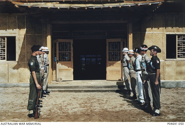 United Nations guards standing at ease outside the hastily built building at Panmunjom, in which the armistice was signed on 27 July 1953. The guard is formed of representatives of some of the major UN nations and includes US Army and Navy military police and unidentified Australian (right) and British (left) soldiers. Two of the men, including the Australian, are wearing the white on red cross-shaped colour patch of the 8th United States Army. All are wearing light blue UN identification tags. 
