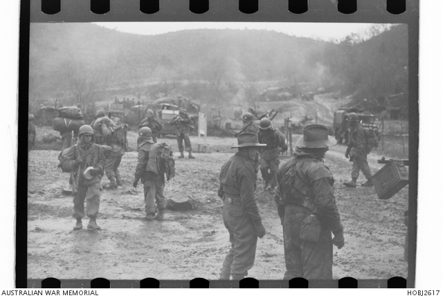 The 3rd Battalion, The Royal Australian Regiment (3RAR), in Korea, is being relieved by troops of the 20th Battalion, Philippines Regimental Combat Team. The Australians have been involved in fighting in the Battle of Maryang San and an area near the Imjin River. In a camp beside a valley, two unidentified soldiers from 3RAR stand in the mud and watch the newly arrived members of the 20th Battalion acclimatise to the camp. Photo circa 23 November 1951