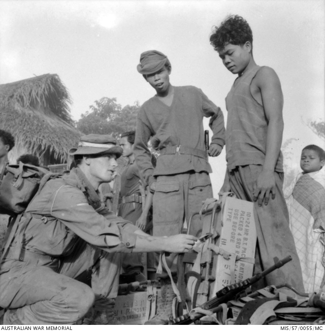 Corporal (Cpl) Jeff Smith, 23, of Launceston, Tasmania, 2nd Battalion, The Royal Australian Regiment (2RAR), demonstrating how to adjust the Alice or 'A' frame which is being used for carrying a 44 pound carton of ration packs. His Malayan bearers, recruited from the Sakai Reserve, listen and watch carefully prior to departing on a re supply mission. Cpl Smith's Owen gun is lying across an Alice frame in the foreground.